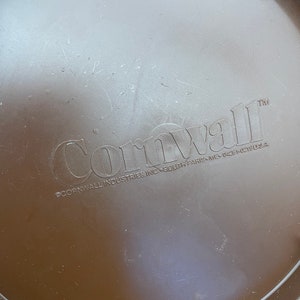 May include: A brown plastic lid with the embossed word "Cornwall" and the text "&copy;Cornwall Industries Inc. South Paris, ME 04281-0319 U.S.A." The lid has a slightly textured surface and is circular in shape.