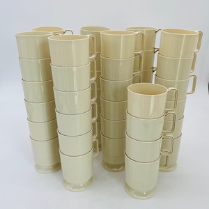May include: A collection of beige-colored mugs with gold-colored handles. The mugs are stacked in multiple columns, showcasing their simple, yet elegant design. The mugs have a pedestal base and a smooth, clean finish.