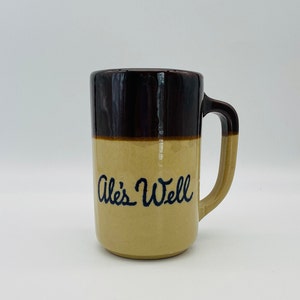 May include: A ceramic mug with a brown top and handle, and a tan base. The words "Ale's Well" are written in blue script on the front. The mug is designed for beverages.
