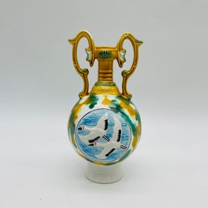 Pode incluir: Um vaso decorativo de cerâmica com base branca, corpo redondo e duas alças. O vaso apresenta um desenho de dois pássaros brancos em voo sobre um fundo azul. O vaso é pintado com detalhes em amarelo, verde e branco.