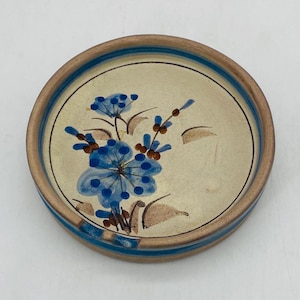 Op de afbeelding: Ronde keramische onderzetter met een bloemenontwerp. De onderzetter heeft een beige midden met een blauwe bloem en bruine accenten. De rand is lichtbruin met twee blauwe strepen. Een decoratief stuk voor in huis.