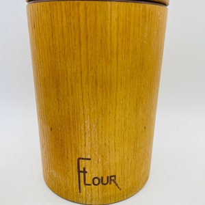 May include: A wooden flour canister with a lid. The cylindrical canister has a light brown wood grain finish. The word "Flour" is printed on the front in a stylized font. The lid is a lighter shade of brown.