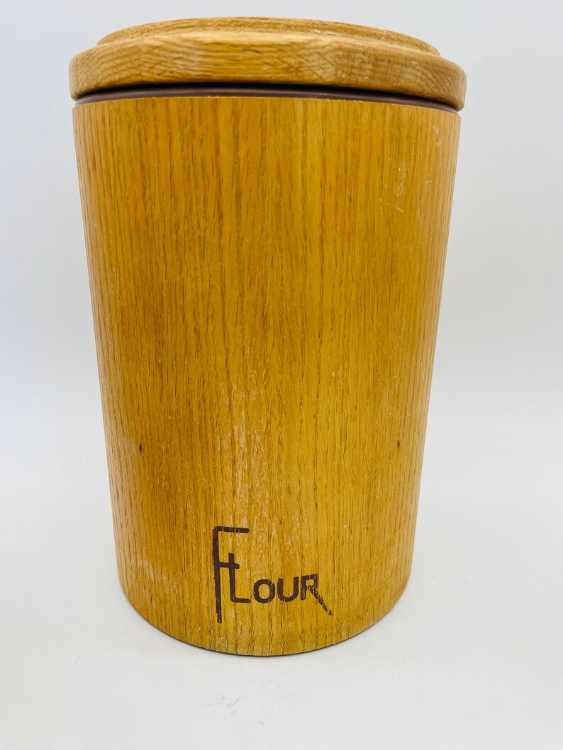 May include: A wooden flour canister with a lid. The cylindrical canister has a light brown wood grain finish. The word "Flour" is printed on the front in a stylized font. The lid is a lighter shade of brown.
