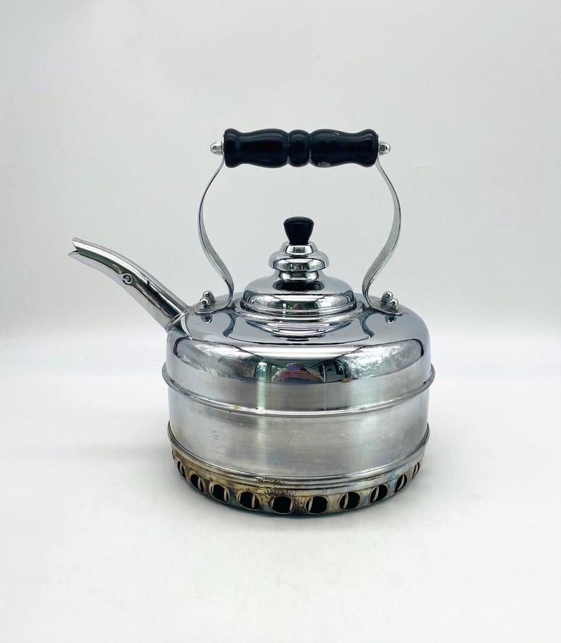 May include: A polished chrome tea kettle featuring a cylindrical body, a curved spout, and a black handle. The lid has a small black knob, and the base is encircled by a row of circular ventilation holes with visible heat discoloration.