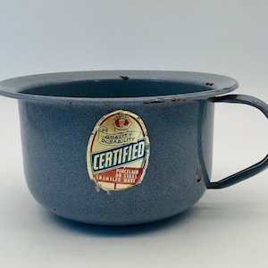 Op de afbeelding: Een vintage blauwe emaille po met een handvat. De pot heeft een brede rand en een vervaagde "Certified" sticker. Het emaille is gespikkeld en vertoont tekenen van slijtage. De pot heeft een lichtblauwe kleur.