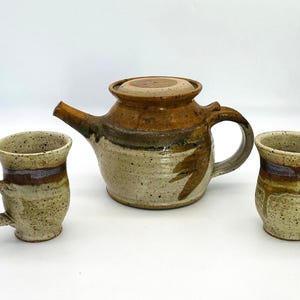 May include: A ceramic teapot with a lid and two matching mugs, all featuring a speckled cream base with brown and purple glazed accents. The teapot has a curved handle and a spout, while each mug has a single handle and a flared rim design.