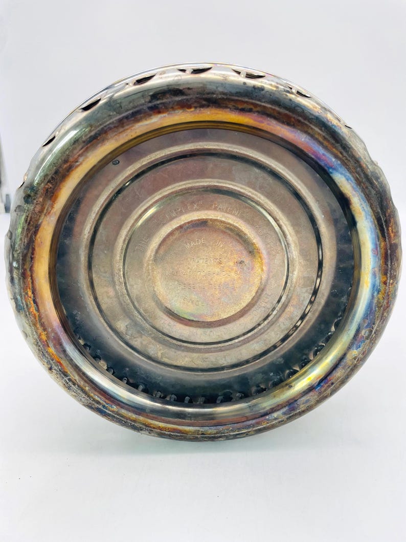 May include: A circular metal object with a tarnished silver-tone finish and a decorative scalloped edge. The centre features embossed text that reads "SIMPLEX PATENT" and "MADE IN ENGLAND," with concentric circular grooves surrounding the base.