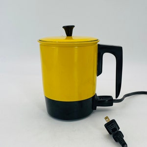 Op de afbeelding: Een vintage gele elektrische koffiepot met een zwarte basis, handvat en dekselknop. Het snoer en de stekker zijn zwart. De koffiepot is ontworpen om koffie te zetten en is waarschijnlijk uit het midden van de 20e eeuw.
