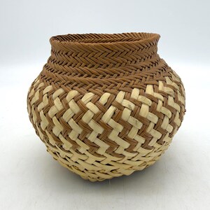 May include: A round, hand-woven basket featuring a wide, open top. The upper section is constructed with a tight, dark brown braided weave, while the lower body displays a contrasting diagonal lattice pattern of light cream and brown strips.