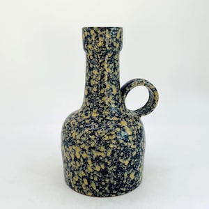 May include: A speckled ceramic vase with a handle. The vase has a dark blue base colour with yellow speckles. The vase has a cylindrical neck and a rounded body. The handle is a circular loop.