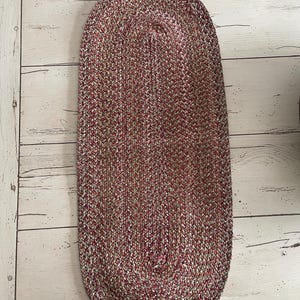 May include: Oval braided rug in shades of red, white, and green. The rug has a textured surface and a braided design. The rug is placed on a light-colored wooden floor. This rug is suitable for a living room or bedroom.