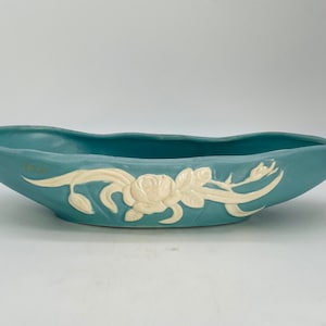 Könnte beinhalten: Eine türkisfarbene, längliche Keramikschale mit einem cremefarbenen floralen Reliefdesign. Die Schale hat eine geschwungene Form und eignet sich zur Präsentation von Pflanzen oder Blumen. Das florale Design umfasst eine Rose und andere botanische Elemente.