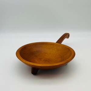 May include: A handcrafted wooden bowl with a handle and three small legs. The bowl is made of light brown wood and has a smooth, polished finish. The handle is curved and has a hole at the end. The bowl is likely used for serving food.