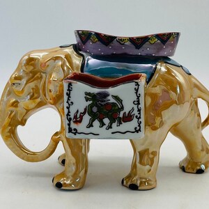 Pode incluir: Estatueta de elefante de cerâmica com acabamento dourado iridescente. O elefante tem uma sela decorativa com um design roxo e azul. Um recipiente retangular branco com uma ilustração verde e vermelha é preso na lateral.