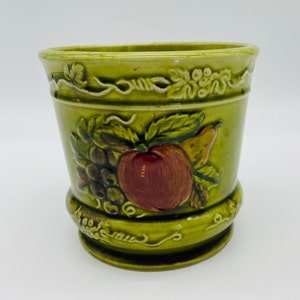 May include: Green ceramic planter with embossed fruit design, including grapes, an apple, and a pear. The planter has a cylindrical shape with a decorative rim and base. The color is a muted green.