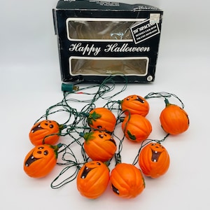 Op de afbeelding: Een slinger van oranje pompoenvormige Halloween-lampjes met lachende gezichten en groene bedrading. De lampjes staan naast een zwarte doos met de tekst "Happy Halloween". De doos heeft een sticker met de tekst "20" afstand tussen elke lamp voor gemakkelijk decoreren.