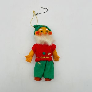 May include: A vintage Christmas ornament in the shape of a gnome. The gnome has a red shirt, green pants, and a green hat. It has a white beard, rosy cheeks, and brown shoes. A black wire hook is attached for hanging.