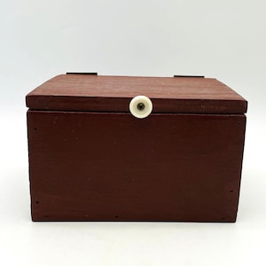 May include: A rectangular wooden box stained in a deep reddish-brown tone. The box features a hinged lid with a small, circular white knob centered on the front edge. Two dark metal hinges are visible along the top rear edge of the container.