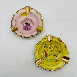May include: Two decorative ashtrays. One is pink with a couple under a tree, the other is yellow with a person on a bridge. Both have gold trim and cigarette rests.