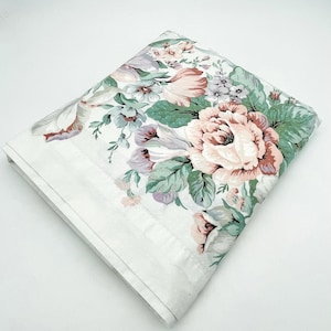 May include: Folded white floral bed sheet featuring large pink, peach, and purple roses with green leaves. The sheet has a white border with a subtle dotted pattern. The overall design is a classic, vintage floral print.