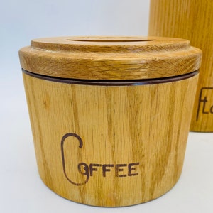 May include: A light brown wooden coffee canister with a matching lid. The word "Coffee" is printed on the front in a dark brown script. The canister has a cylindrical shape. The canister is made of wood.