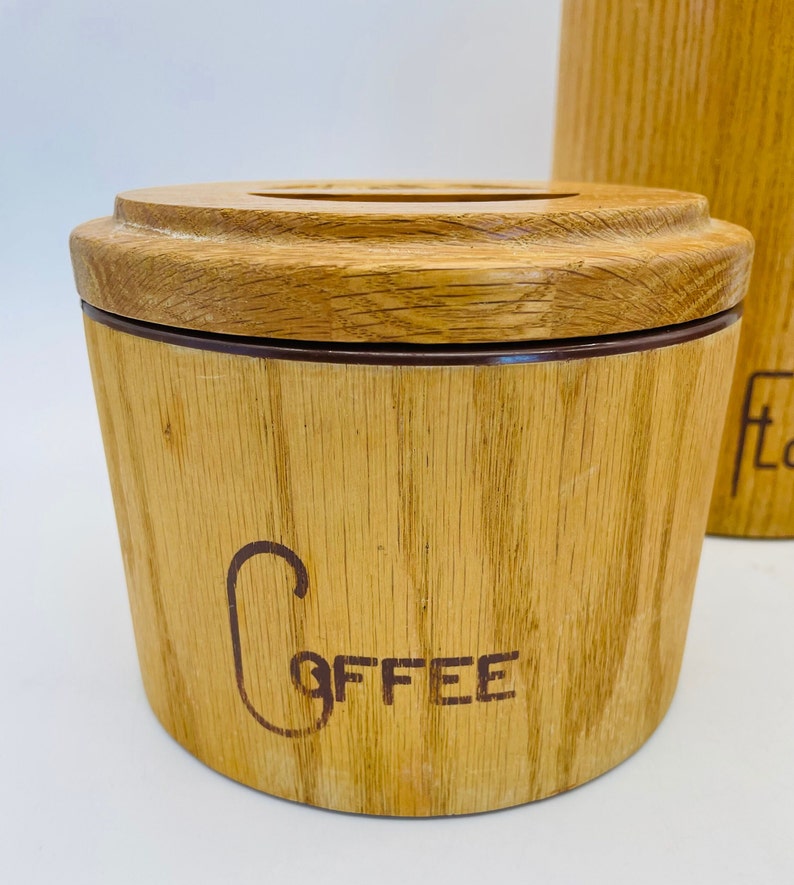 May include: A light brown wooden coffee canister with a matching lid. The word "Coffee" is printed on the front in a dark brown script. The canister has a cylindrical shape. The canister is made of wood.
