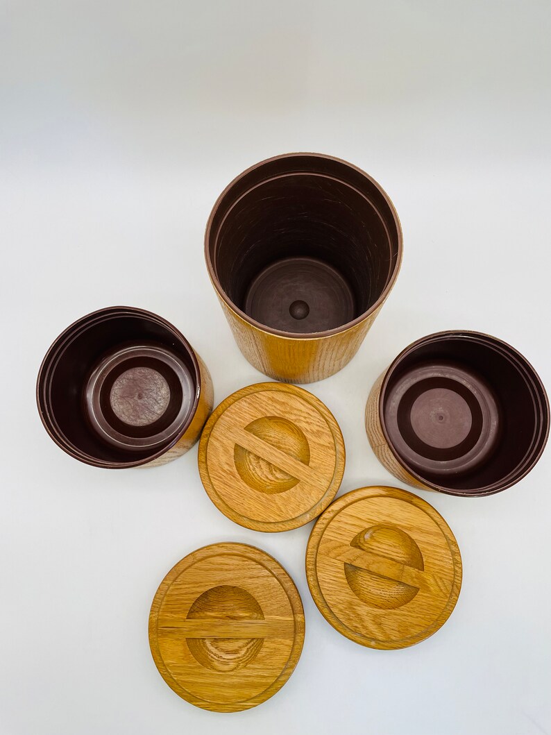 May include: A collection of three brown and tan plant pots with matching wooden lids. The pots are cylindrical with a textured exterior. The lids are round with a carved design. The interior of the pots is a dark brown colour.