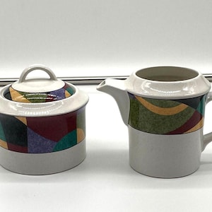 Peut inclure: Un sucrier en céramique avec couvercle et un pot à crème. Le sucrier et le pot à crème ont une base blanche et sont décorés de motifs géométriques colorés verts, rouges et violets.