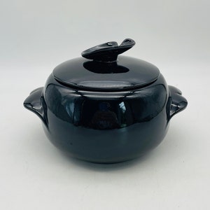 May include: A glossy black ceramic casserole dish with a lid. The lid has a decorative handle. The dish has two handles on the sides. The casserole dish is round and has a smooth, reflective surface.