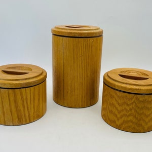 May include: Three wooden containers of varying sizes. The containers are cylindrical with a light brown finish and a dark brown band near the base. Each has a lid with a slot. The wood grain is visible.