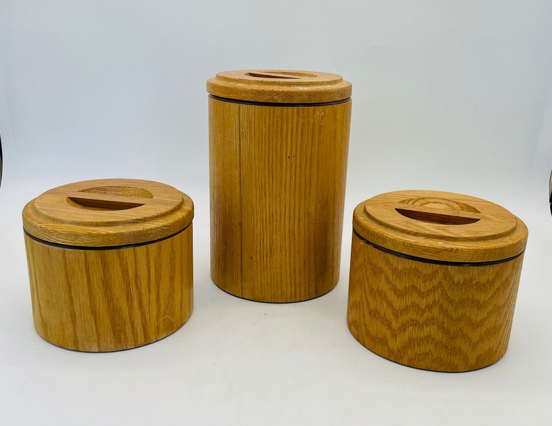 May include: Three wooden containers of varying sizes. The containers are cylindrical with a light brown finish and a dark brown band near the base. Each has a lid with a slot. The wood grain is visible.