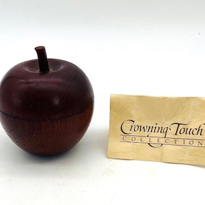 May include: A wooden apple-shaped box with a dark red top and a lighter brown base. The box has a small, dark brown stem. A small, gold-colored tag with the text "Crowning Touch Collection" is next to the box.