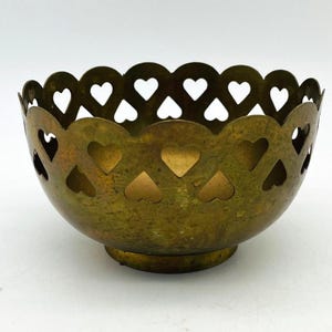 May include: A vintage brass bowl featuring heart-shaped cutouts. The bowl has a scalloped rim and a circular base. The brass has a tarnished, aged appearance. The bowl is likely used for decorative purposes or as a small container.