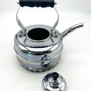 May include: A chrome-finished metal kettle with a black handle and a matching removable lid. The kettle features a wide, circular body with a short, angled spout and a perforated base. The lid has a small black knob on top for easy handling.