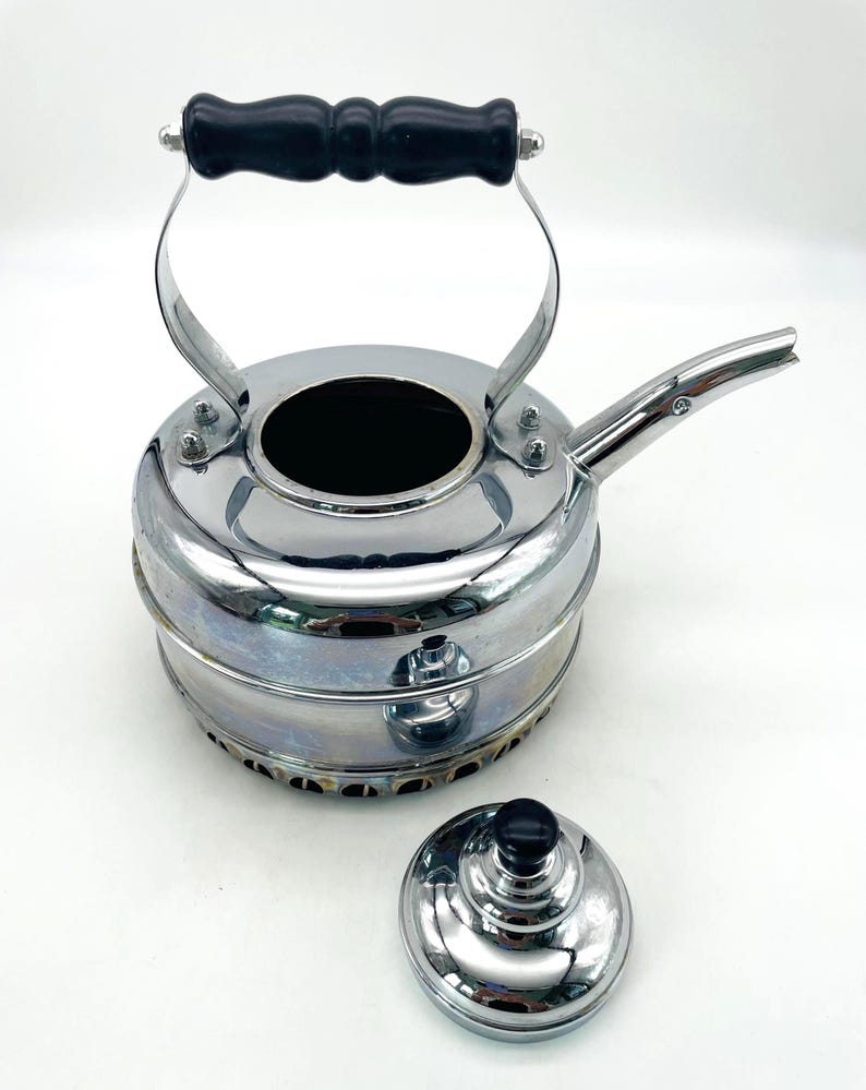 May include: A chrome-finished metal kettle with a black handle and a matching removable lid. The kettle features a wide, circular body with a short, angled spout and a perforated base. The lid has a small black knob on top for easy handling.