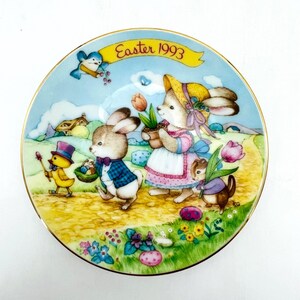 Könnte beinhalten: Ein runder dekorativer Porzellanteller mit Goldrand, der eine bunte Illustration von anthropomorphen Kaninchen und einem Küken auf einem Weg zeigt. Der Text "Easter 1993" steht auf einem gelben Banner oben. Die Szene umfasst Blumen, einen blauen Himmel und ein kleines Haus im Hintergrund.