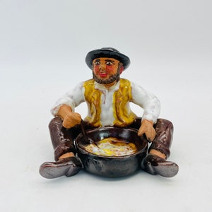 Könnte beinhalten: Keramikfigur eines sitzenden Mannes, der in einem Topf rührt. Der Mann trägt einen schwarzen Hut, ein weißes Hemd, eine gelbe Weste und eine braune Hose. Der Topf ist dunkelbraun und enthält eine cremige, gelbe Mischung. Auf weißem Hintergrund.