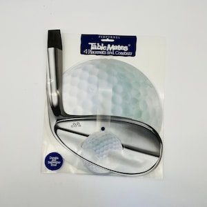 May include: A set of Table Mates placemats and coasters featuring a golf theme. The design includes a golf club and a golf ball. The packaging includes the text "Durable and Dishwasher Proof".
