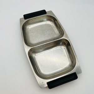 Könnte beinhalten: Ein zweiteiliges Serviertablett aus Edelstahl. Das Tablett hat eine rechteckige Form mit abgerundeten Ecken und schwarzen Griffen an jedem Ende. Das Innere des Tabletts hat eine gebürstete Metalloberfläche.