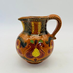 May include: A small, ceramic pitcher with a handle and spout. The pitcher is decorated with a brown, orange, and yellow floral design. The handle and spout are brown. The pitcher is likely used for serving liquids.
