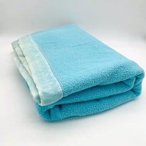 May include: A folded light blue blanket with a textured, fuzzy surface and a smooth, lighter blue satin-like border trim. The blanket is neatly stacked, showcasing the soft material and the contrast between the fleece-like body and the trim.