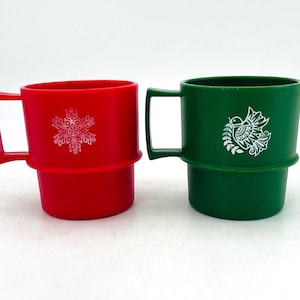 May include: A red mug with a white snowflake graphic and a green mug with a white bird graphic. Both mugs feature a square handle, a cylindrical body with a raised horizontal ridge around the center, and a smooth, matte finish on the exterior.