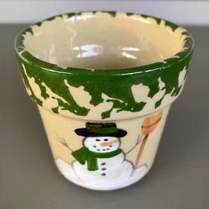 May include: A small, hand-painted ceramic flower pot featuring a snowman design. The pot has a cream-colored base with a green rim and a painted snowman wearing a black hat, green scarf, and holding a broom. The pot is approximately 4 inches tall.