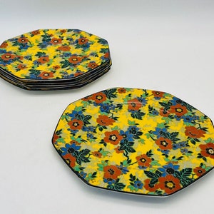 May include: A set of five octagonal ceramic plates featuring a vibrant yellow background with a dense floral pattern of red, blue, and green blossoms and leaves. Each plate has a thin black rim, and they are arranged with one plate flat.