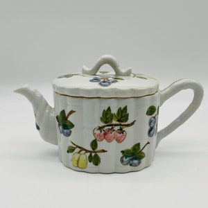 May include: White ceramic teapot with a lid, decorated with hand-painted fruit and leaf designs. The teapot features a curved handle and spout, and the lid has a small loop for easy removal. The fruit includes strawberries, blueberries, and yellow plums.