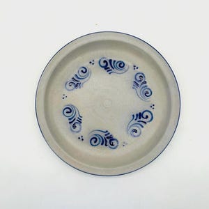 May include: A round, beige ceramic plate with a dark blue rim and decorative blue scrollwork around the center. The plate has a simple, yet elegant design, suitable for everyday use or special occasions.