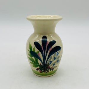 Può includere: Un piccolo vaso in ceramica color crema con la parte superiore svasata e il corpo arrotondato. Il vaso è decorato con disegni floreali dipinti a mano nei toni del blu, verde e rosa. Una fascia verde circonda la base.