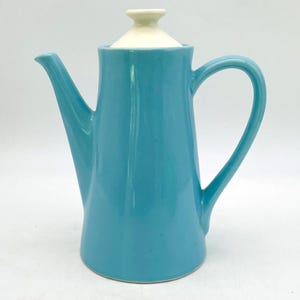 May include: A bright turquoise ceramic coffee pot with a smooth, glossy finish. The vessel features a tall, tapered body, a curved handle on the right side, a long spout on the left, and a removable, conical off-white lid with a small knob top.