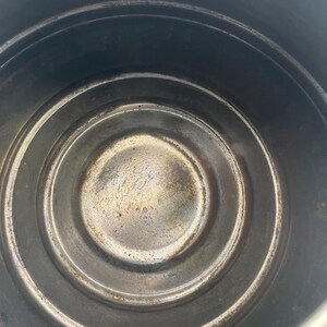 May include: A top-down view of the interior of a circular metal pot or container. The base features concentric circular ridges and shows signs of wear, discolouration, and dark metallic staining across the bottom surface and the vertical walls.