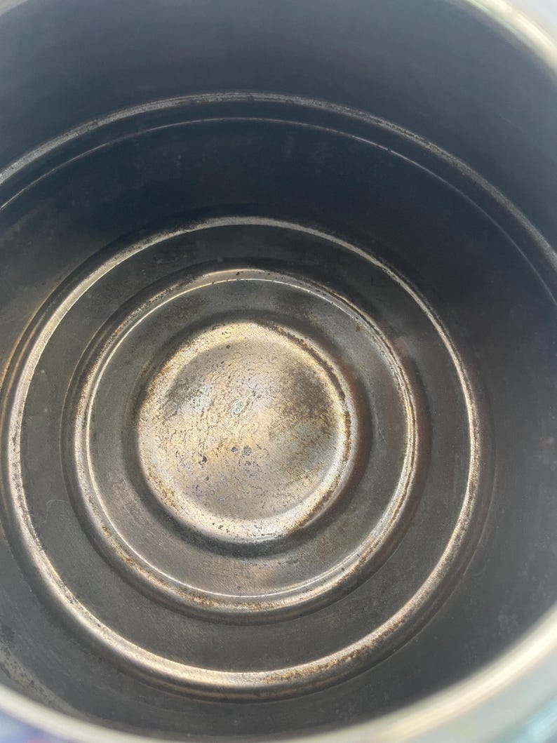 May include: A top-down view of the interior of a circular metal pot or container. The base features concentric circular ridges and shows signs of wear, discolouration, and dark metallic staining across the bottom surface and the vertical walls.